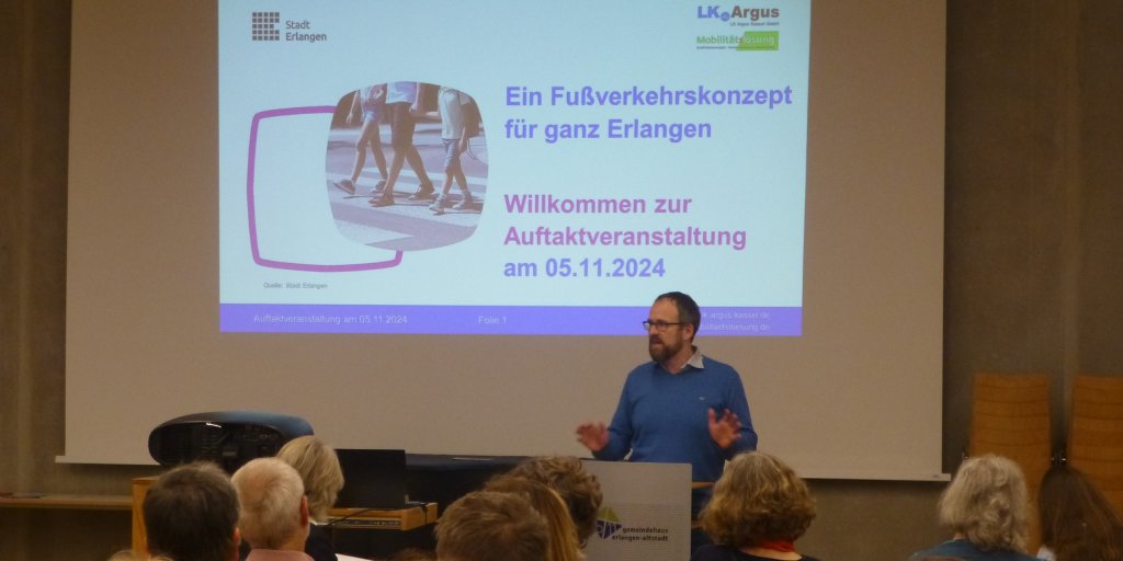 Foto mit einer Leinwand im Hintergrund, vor der ein Mann eine Rede hält.