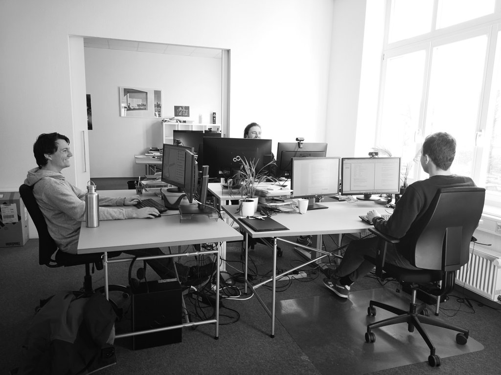 Foto von drei jungen Männern, die an Schreibtischen vor ihren Monitoren sitzen und sich anlächeln.