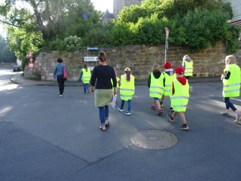 Foto mit mehreren Kindern in Warnweste sowie zwei Frauen, die alle eine Straße queren.