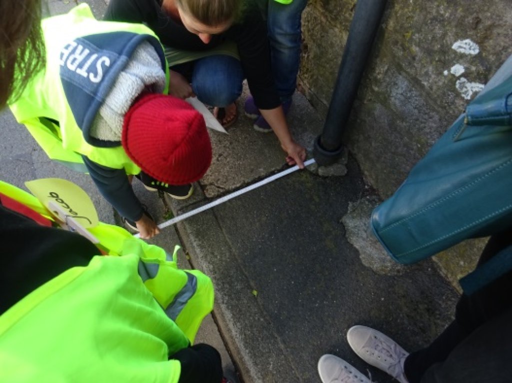 Foto mit einem Kind in Warnweste, das einen sehr schmalen Gehweg ausmisst, und weiteren Personen außenrum.