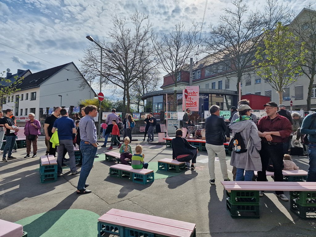 Foto eines Platzbereichs im Straßenraum mit Bänken in Form von rosa Brettern auf grünen Kisten sowie mit mehreren sich unterhaltenden Menschen.